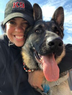 Jessie with a german shepherd who has their tongue out, Jessie is wearing a PrairieBurn K9 Academy Hat